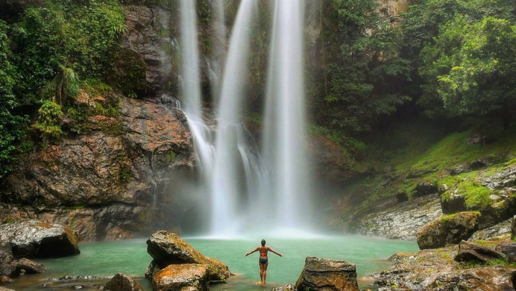 person standing in front of cunca rami waterfall surrounded by lush forest