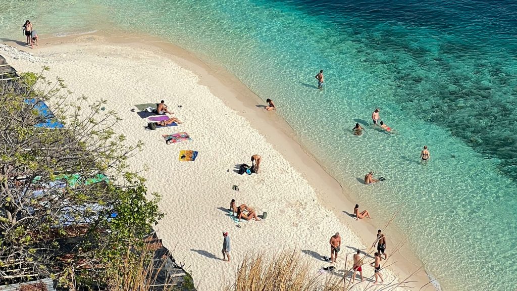 Tourists swimming and relaxing on white sand beach at Kelor Island with clear shallow water