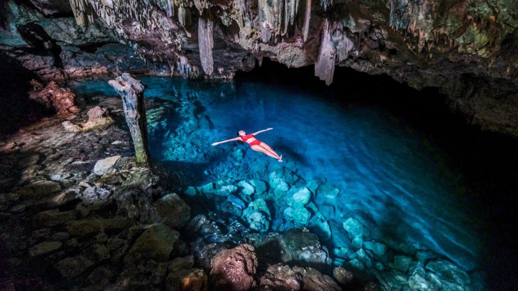 person floating in a clear blue underground cave pool surrounded by stalactites